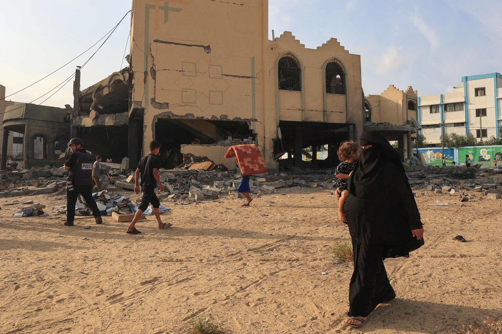 Um repórter fotográfico e civis passam por um prédio danificado em Khan Yunis, após ataques aéreos israelenses no sul da Faixa de Gaza — Foto: SAID KHATIB/AFP