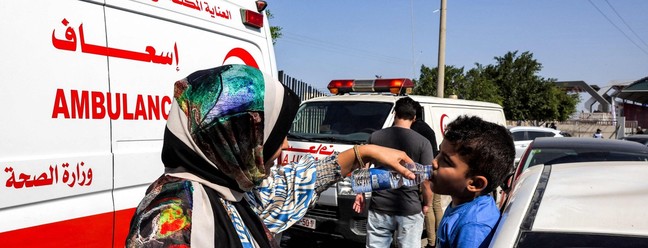 Uma mulher ajuda uma criança a beber água de uma garrafa enquanto espera ao lado das ambulâncias do Ministério da Saúde palestino — Foto: Mohammed ABED/AFP