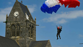 Um paraquedista do Comando da Marinha Francesa guia seu paraquedas vermelho, branco e azul - bandeira da frança - pela igreja de Sainte-Mere-Eglise, como parte das comemorações do "Dia D" que marcam o 80º aniversário do desembarque dos Aliados na Segunda Guerra Mundial na Normandia. — Foto: MIGUEL MEDINA / AFP