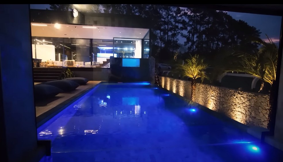 Casa de Boca Rosa à noite: piscina interna na sala, com transparência em vidro e piscina externa, com iluminação especial — Foto: Reprodução/YouTube