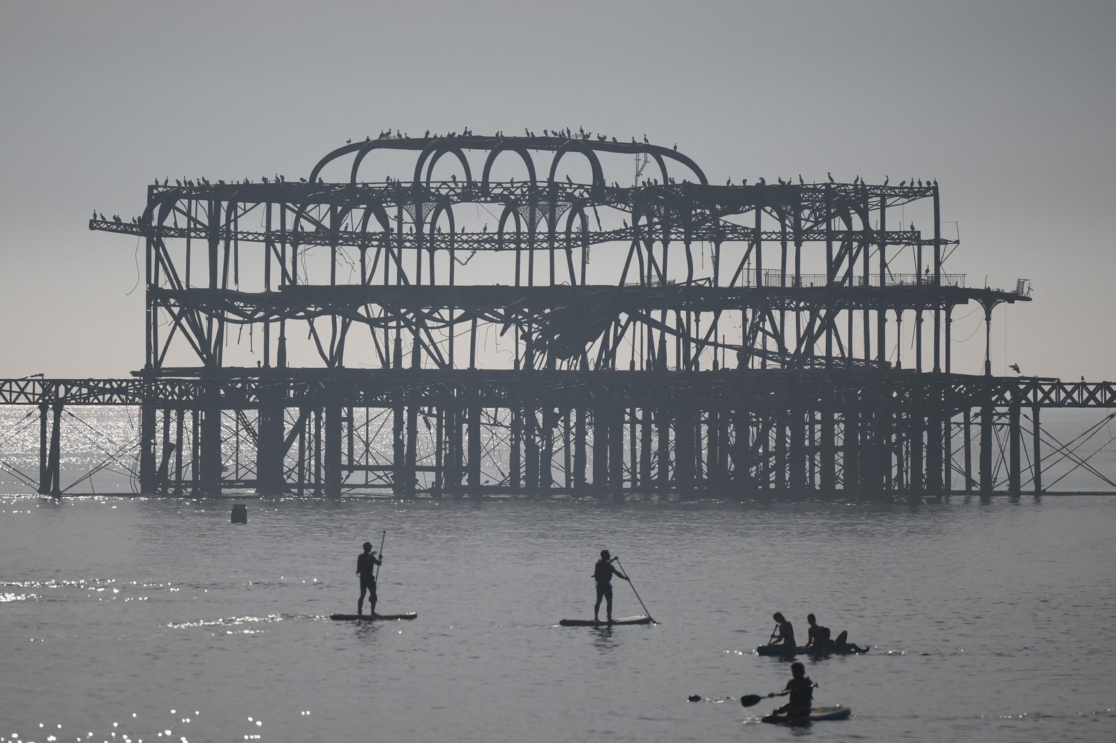 Pessoas praticam paddle e canoagem passando pelo abandonado cais oeste, aproveitando o sol e o mar na praia de Brighton, — Foto: AFP