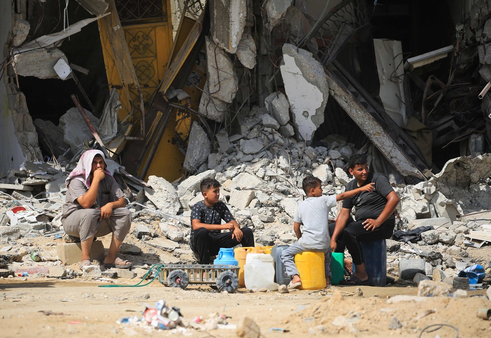 Crianças palestinas sentadas em contêineres de água em frente a edifícios danificados durante bombardeios israelenses anteriores — Foto: Eyad BABA / AFP