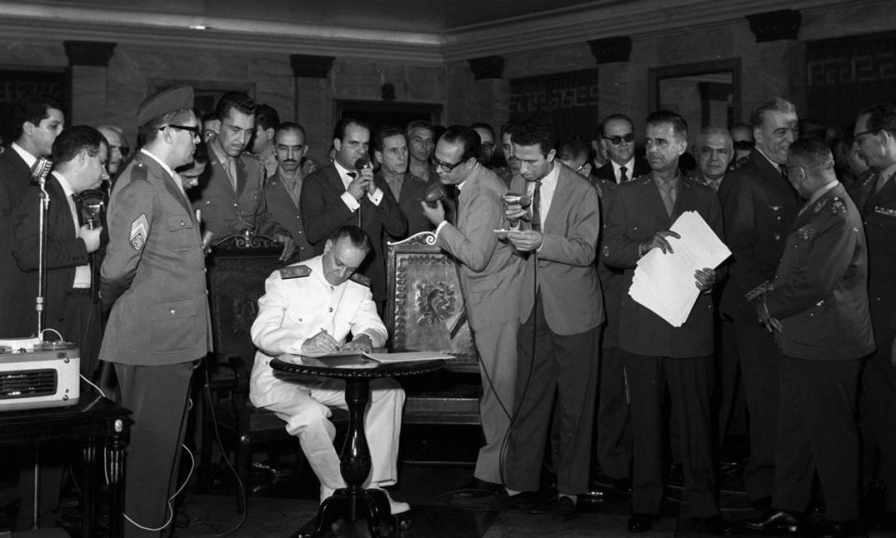 Uma vez deposto João Goulart, os chefes militares passam a emitir decretos, os chamados Atos Institucionais, ignorando a existência do poder Legislativo e da Constituição. Na foto, o ministro da Marinha, Augusto Rademaker, assina o primeiro ato do regime militar, em 9 de abril, aumentando os poderes do presidente da República, que passa então a poder cassar mandatos de parlamentares e suspender direitos políticos — Foto: Agência O Globo