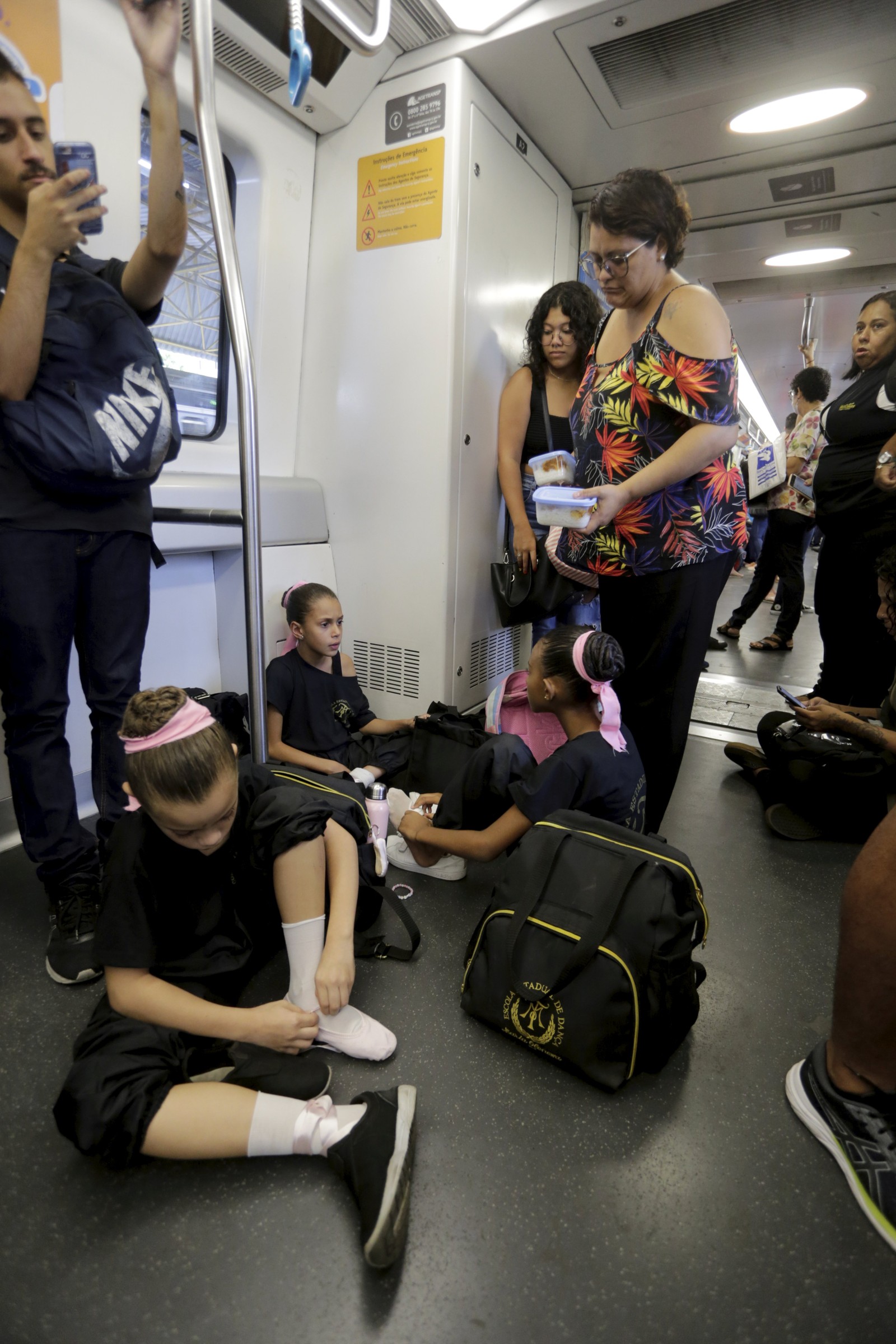Preparativos. Assim que chegam ao metrô, as meninas sentam no canto do vagão para terminarem de se arrumar. No primeiro plano, Eloá calça sapatilha de balé