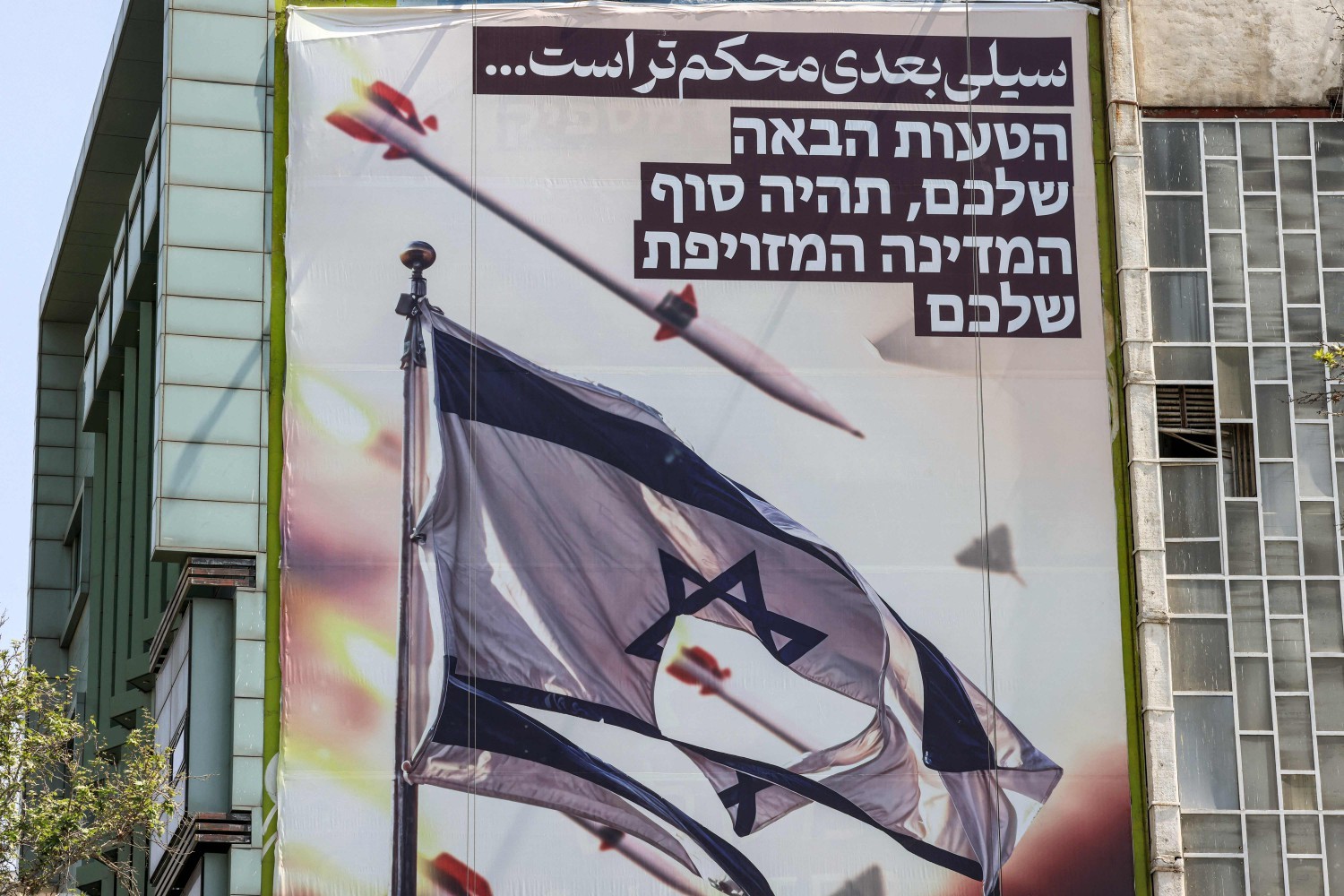 Painel ilustrado na Praça Palestina, em Teerã, mostra mísseis e drones voando perto de uma bandeira israelense rasgada, dizendo, em farsi, "o próximo tapa será mais violento" e, em hebraico, "seu próximo erro será o fim de seu falso Estado" — Foto: ATTA KENARE / AFP