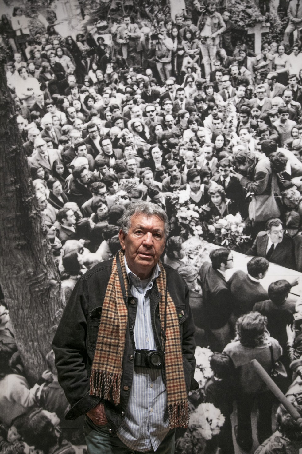 Evandro Teixeira posa na frente de uma das fotos mais emblemáticas da sua carreira, quando registrou o enterro de Pablo Neruda, que se transformou na 1ª grande manifestação contra o regime ditatorial de Augusto Pinochet — Foto: Guito Moreto/Agência O Globo