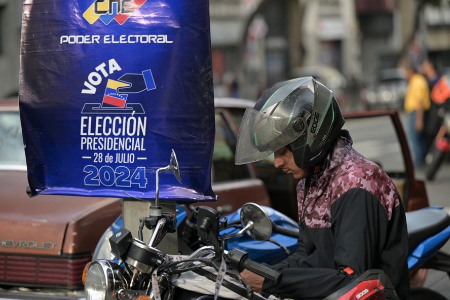 Um motociclista olha seu celular em frente a um cartaz do Conselho Nacional Eleitoral (CNE) promovendo as próximas eleições presidenciais em Caracas, em 23 de julho de 2024