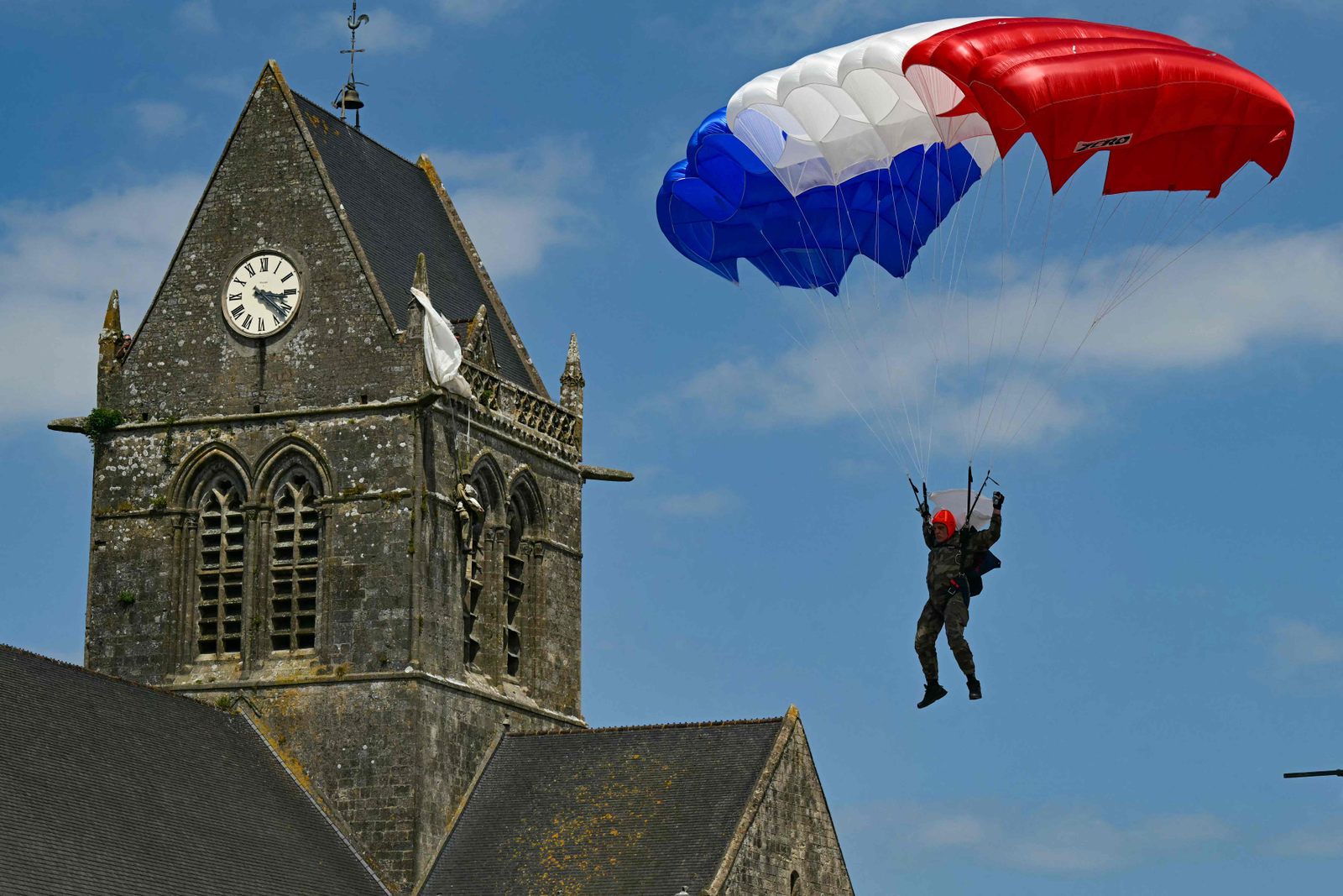 Um paraquedista do Comando da Marinha Francesa guia seu paraquedas vermelho, branco e azul - bandeira da frança - pela igreja de Sainte-Mere-Eglise, como parte das comemorações do "Dia D" que marcam o 80º aniversário do desembarque dos Aliados na Segunda Guerra Mundial na Normandia. — Foto: MIGUEL MEDINA / AFP