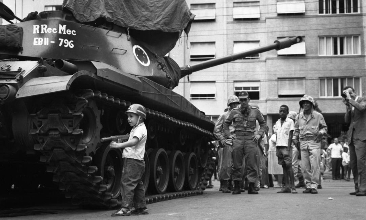 Uma criança usando capacete e portando uma arma de brinquedo é observada por soldados e civis enquanto brinca ao lado de um tanque do Exército — Foto: Agência O Globo