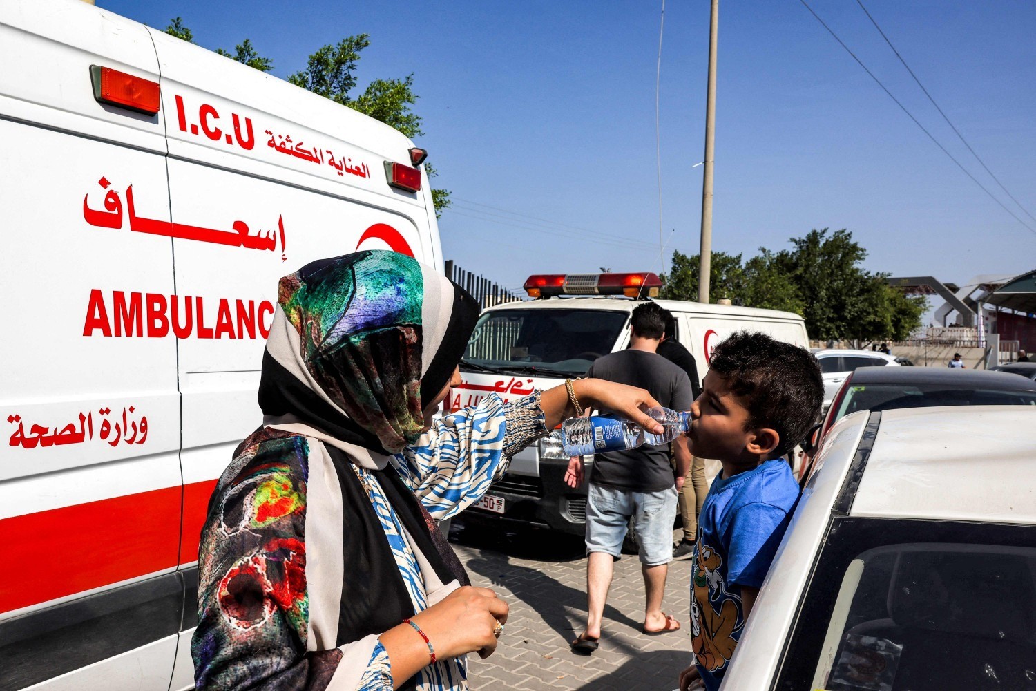 Uma mulher ajuda uma criança a beber água de uma garrafa enquanto espera ao lado das ambulâncias do Ministério da Saúde palestino — Foto: Mohammed ABED/AFP