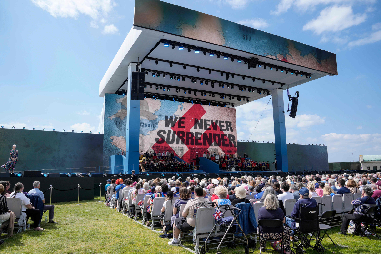 Os convidados assistem a uma banda tocar durante o evento comemorativo nacional do Reino Unido para marcar as comemorações do 80º aniversário do Dia D. Na tela, escrito em inglês, a frase "nós nunca nos rendemos". — Foto: Kin Cheung / POOL / AFP