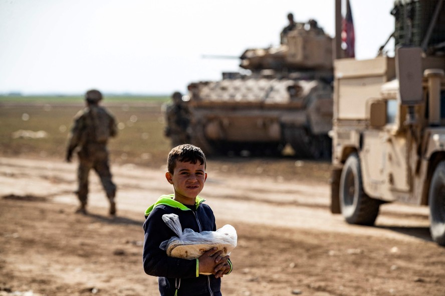 Criança carregando uma sacola com pão ao lado de tropas americanas na cidade de Tal Hamis, na Síria