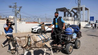 Um jornalista filma com uma câmera de vídeo enquanto um homem conduz uma carroça puxada por um burro carregando bagagens fora da passagem de fronteira de Rafah com o Egito, no sul da Faixa de Gaza — Foto: MOHAMMED ABED / AFP