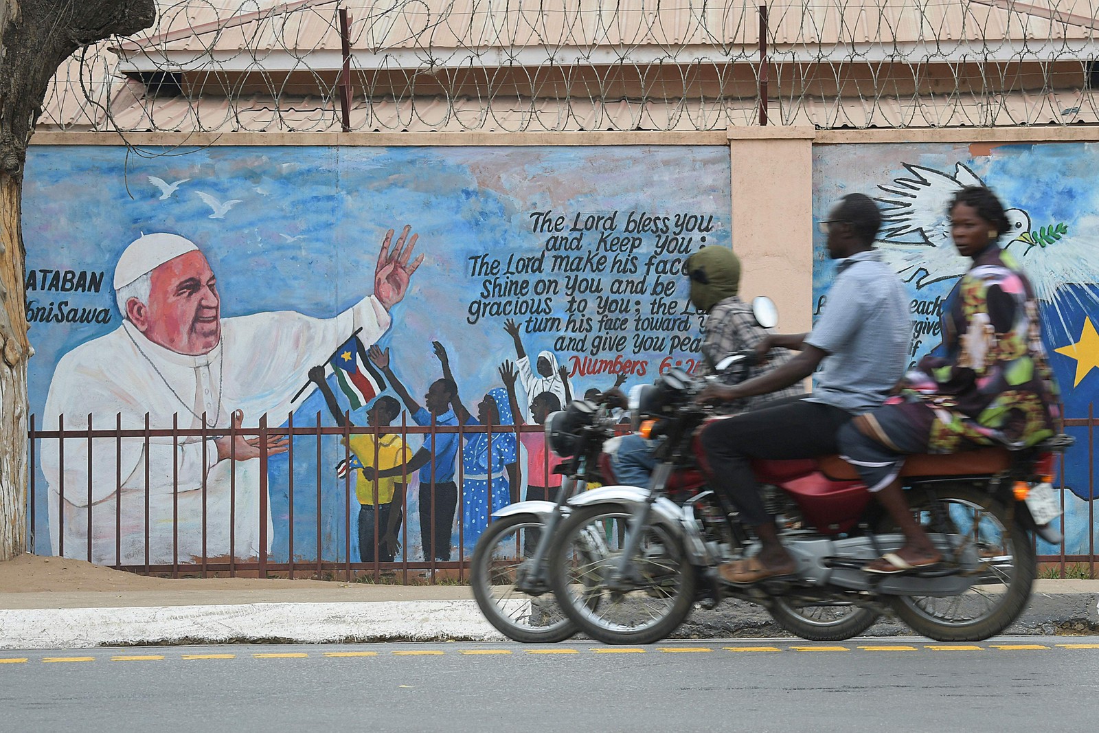 Pessoas em motocicletas passam por um mural representando o Papa Francisco enquanto os preparativos continuam antes de sua visita a Juba, no Sudão do Sul — Foto: SIMON MAINA/AFP