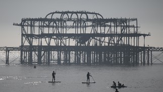 Pessoas praticam paddle e canoagem passando pelo abandonado cais oeste, aproveitando o sol e o mar na praia de Brighton, — Foto: AFP