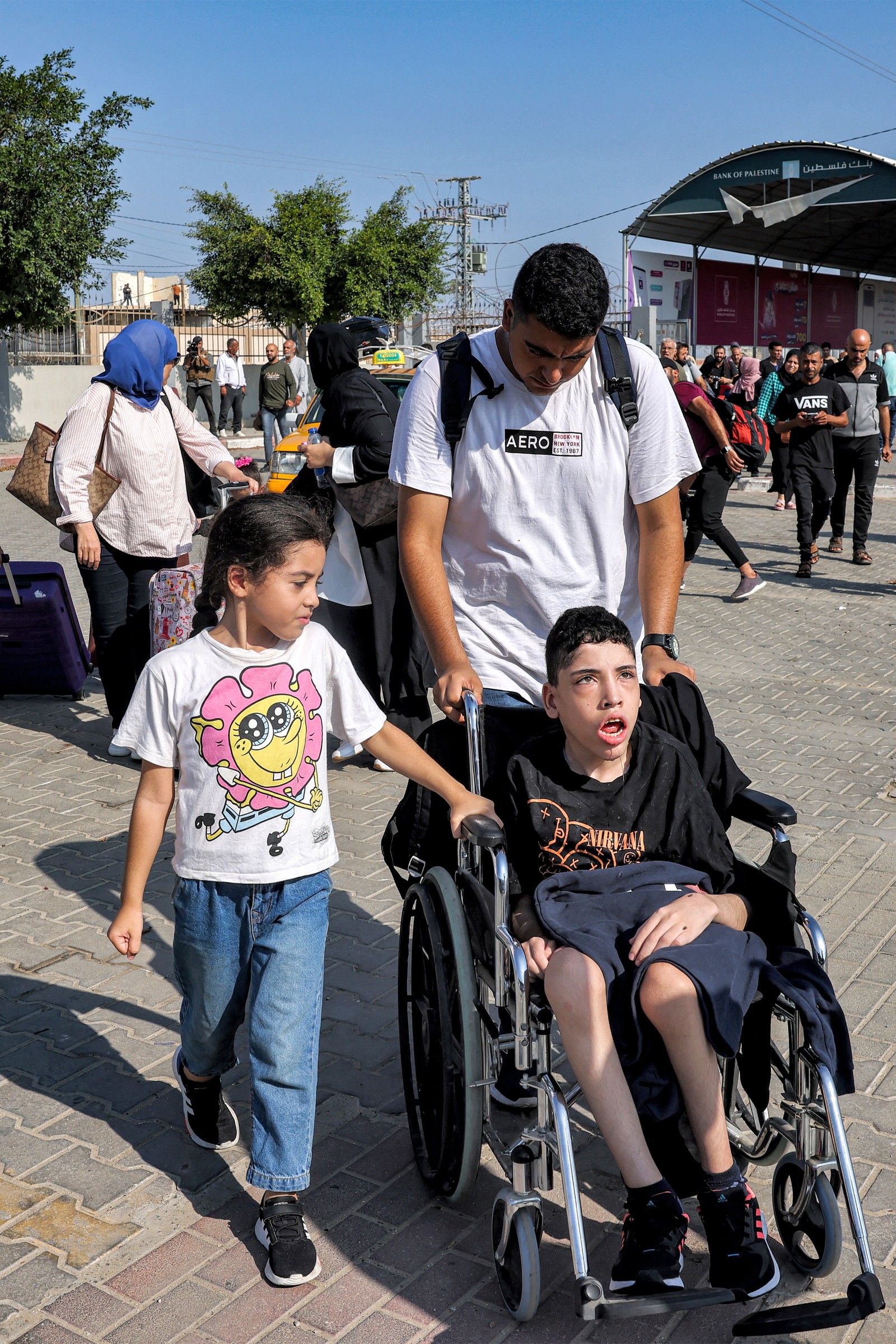 Um homem empurra uma criança numa cadeira de rodas enquanto as pessoas entram na passagem da fronteira de Rafah, no sul da Faixa de Gaza, antes de cruzarem para o Egito — Foto: Mohammed ABED/AFP