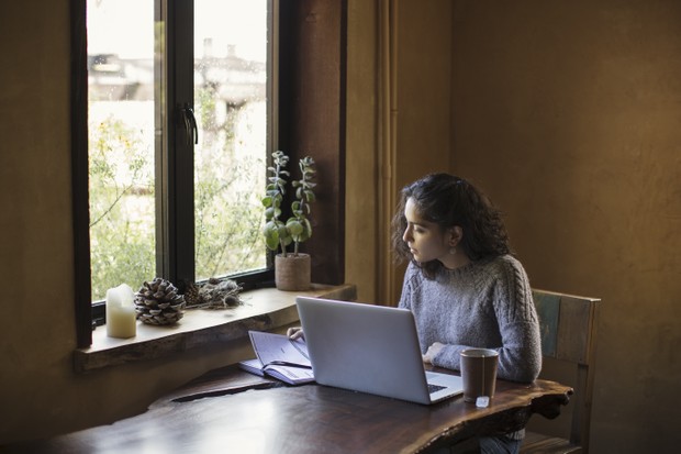 Sitting next to a window, embraced by natural light, this determined young adult (mixed-race) woman is working from home on a tight deadline. She is leaning in and focused to finish her project. Prominent laptop computer, a reference book, and mug of tea  (Foto: Getty Images)