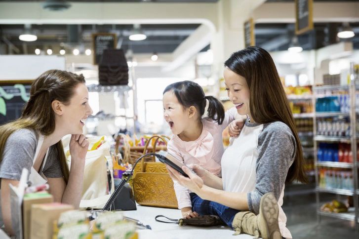 A mother holding her young daughter, conversing with a store clerk at a retail checkout.