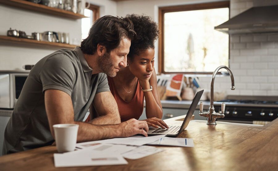 A couple looks at their laptop computer screen at their kitchen as documents are scattered on the table.