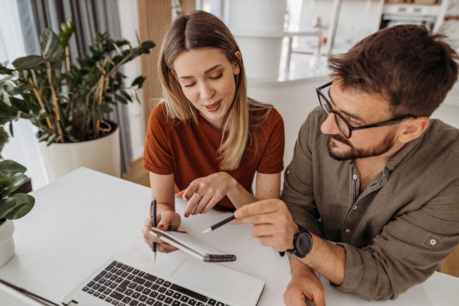 A couple talk to each other as they point at a notepad with next to their laptop.