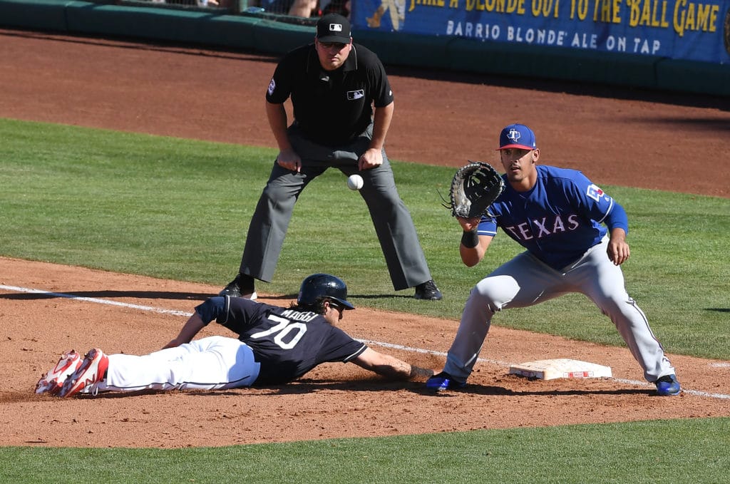 GOODYEAR, AZ - MARCH 02:  Drew Maggi #70 of the Cleveland Indians dives back to first base as Ronald Guzman #67 of the Texas Rangers waits for the throw from the pitchers mound as first base umpire Ed Hickox #15 looks on during the sixth inning of a spring training game at Goodyear Ballpark on March 2, 2018 in Goodyear, Arizona. Chisenhall popped out to the catcher.  (Photo by Norm Hall/Getty Images)