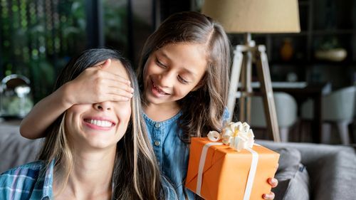 A smiling young girl, holding a box wrapped in orange paper with a white ribbon and bow, covers her mother's eyes to surprise her