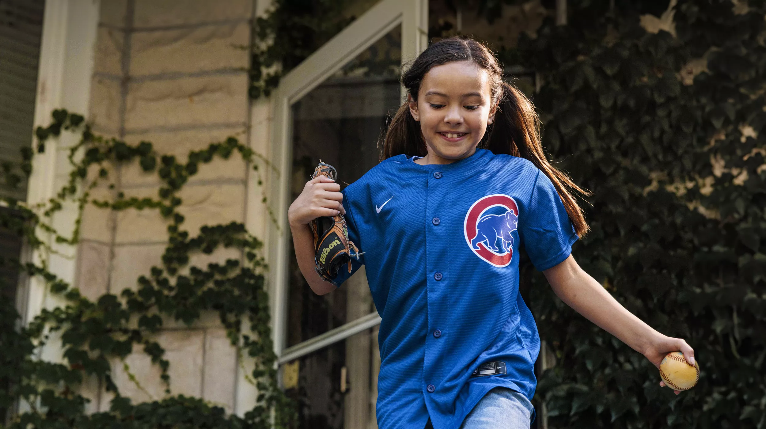 Young girl wearing baseball jersey running outside the front door to play baseball.