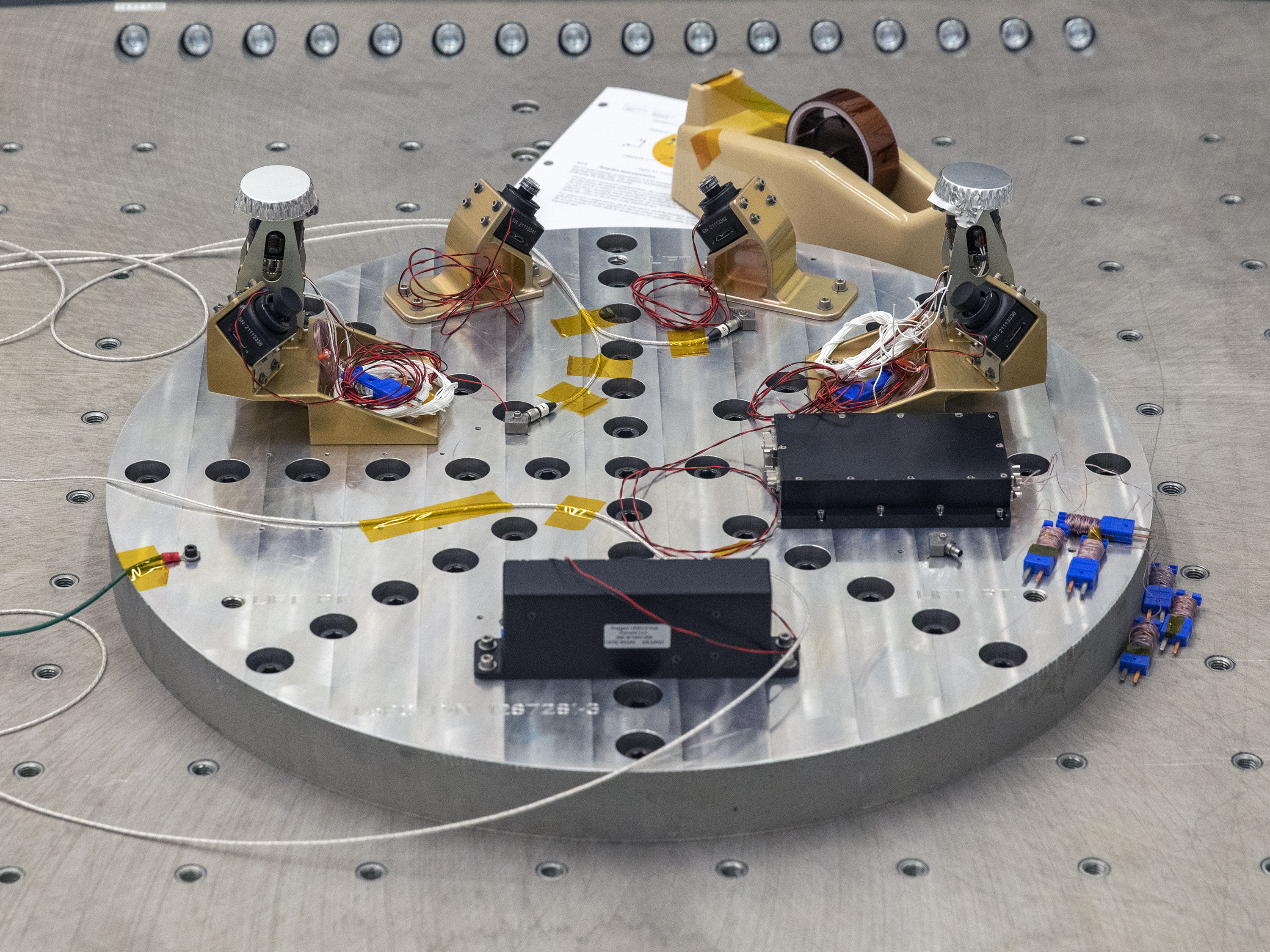 A set of four cameras mounted on a metal wheel, along with control circuitry and processing units, undergo vibration testing in a lab. For some reason, there is a tape dispenser that appears to be from the 70s in the background. I imagine this wasn't part of the experiment.