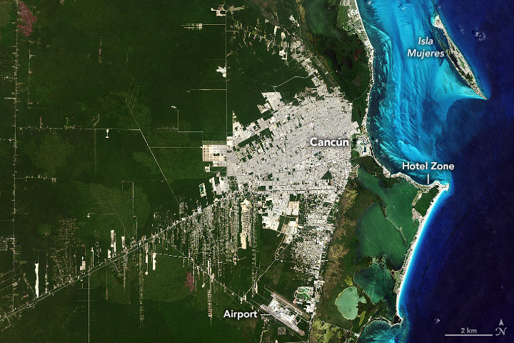 The vast green landspace around the coastal city takes a vast majority of the image space, with the water to the right appearing in a gradient of vibrant teal to a deep dark blue farther from the coast. The human settlement is centered primarily around a single road in until you near the coast, at which point the city is densely packed and reflects bright white with only a few, and mostly small green spaces interspersed.