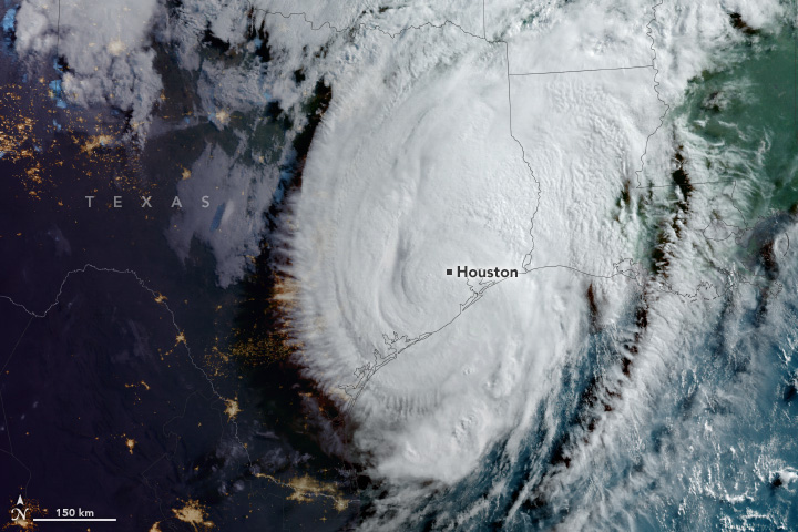 A circular cloud formation is shown with the coastal outline of Southern Texas and Louisiana superimposed on the image. The cloud centers on Houston and at the edges of the blanket of white clouds radiate outward in spokes along the left of the storm. Western Texas city lighting can also be seen in the dark.