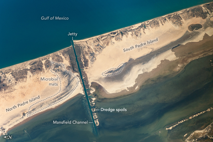 A barrier island spans the image diagonally, with the Gulf of Mexico at the top and inland water at the bottom. The strip of land is almost entirely beige sand, however the Gulf side has some vegetation creating dark patches and affecting the topography as well as a patchy dark shade sweeping through the more central part of the island. Splitting the island is a dredged channel and jetty with the dredged sand being left in the inland waterway to create small islands of its own.