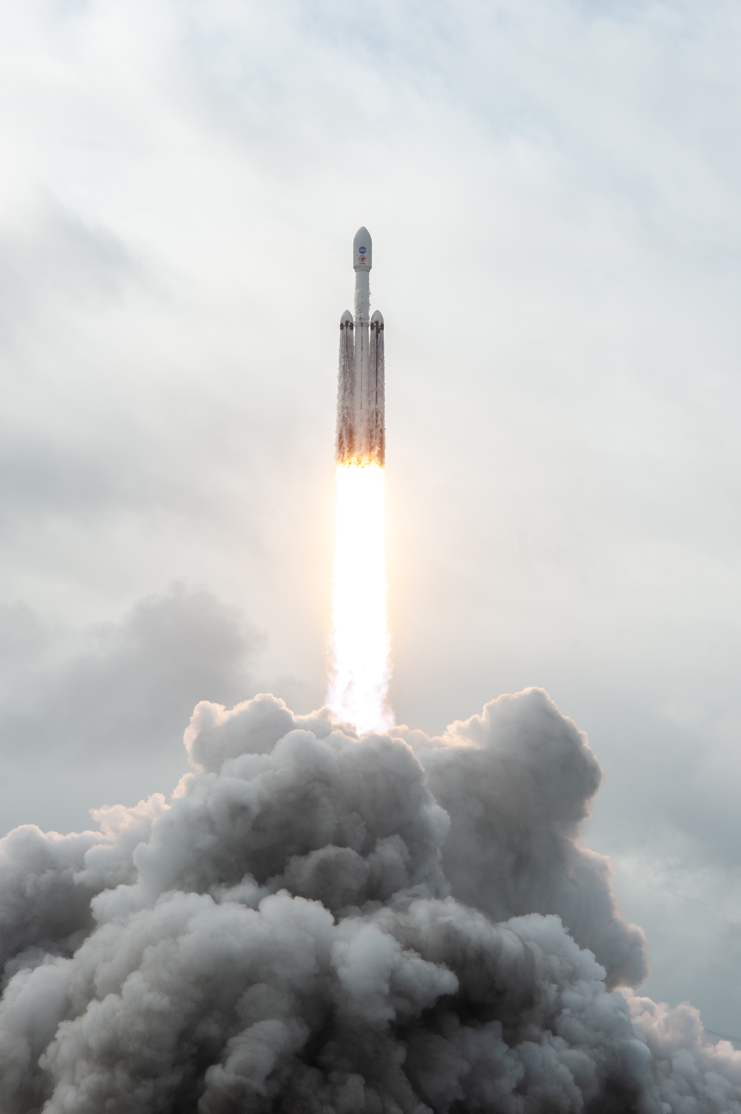 The Psyche spacecraft rides to space inside its faring on top of a rocket with two side boosters. The spacecraft faring and boosters are all white. Flames shoot out the bottom of the rocket stack, and clouds are beneath the flames.