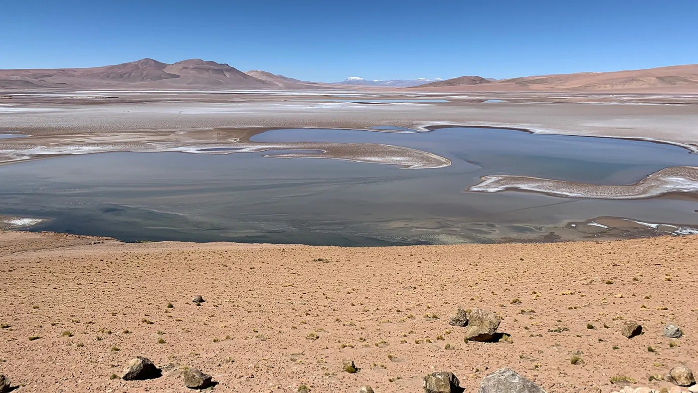 A landscape scene looks out on a mostly flat surface with pools of liquid. in the middle A mountain range in the distance runs across the top of the image, punctuated by a daylight blue sky.