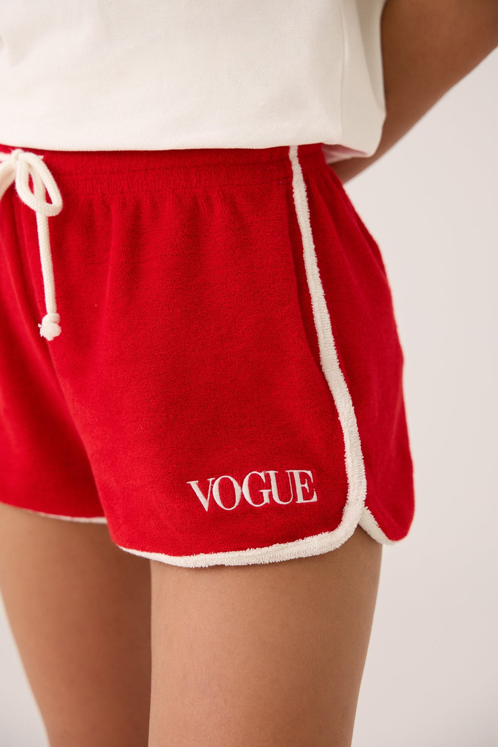 Model seitlich stehend in VOGUE Shorts Rot mit Logo-Stickerei: Detailansicht VOGUE Shorts Rot mit Logo-Stickereiei kombiniert mit VOGUE Polo-Shirt Weiß mit Logo-Stickerei