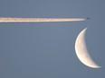 A crescent moon is in the bottom right hand of the frame against a blue sky. Above it, there's a plane leaving a contrail behind it that crosses the entire photo to the left side.