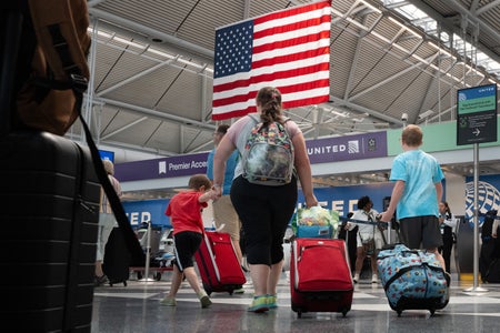 Passengers walk through an airport pulling suitcases with an American flag hanging from the ceiling