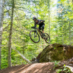 Mountain biker going off a jump on a dirt track with trees in the background. 