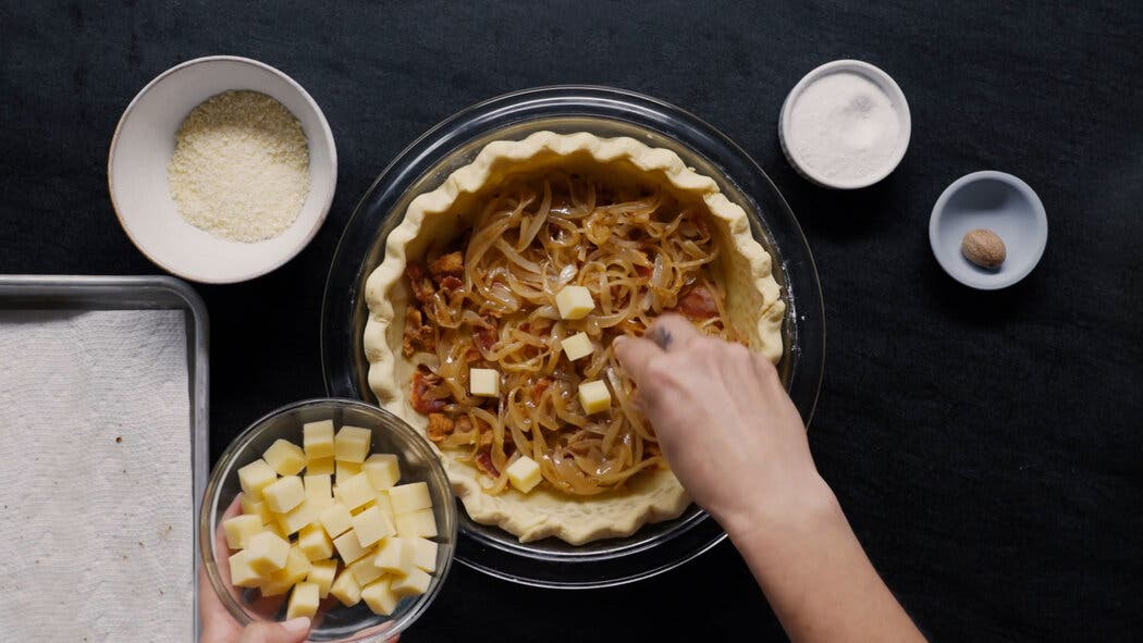 Image of bacon, onions and cheese being place in a par-baked pie crust for making a quiche Lorraine. 