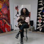 A woman sits cross-legged on a stool holding a cat with two large, colorful paintings propped on the walls behind her.