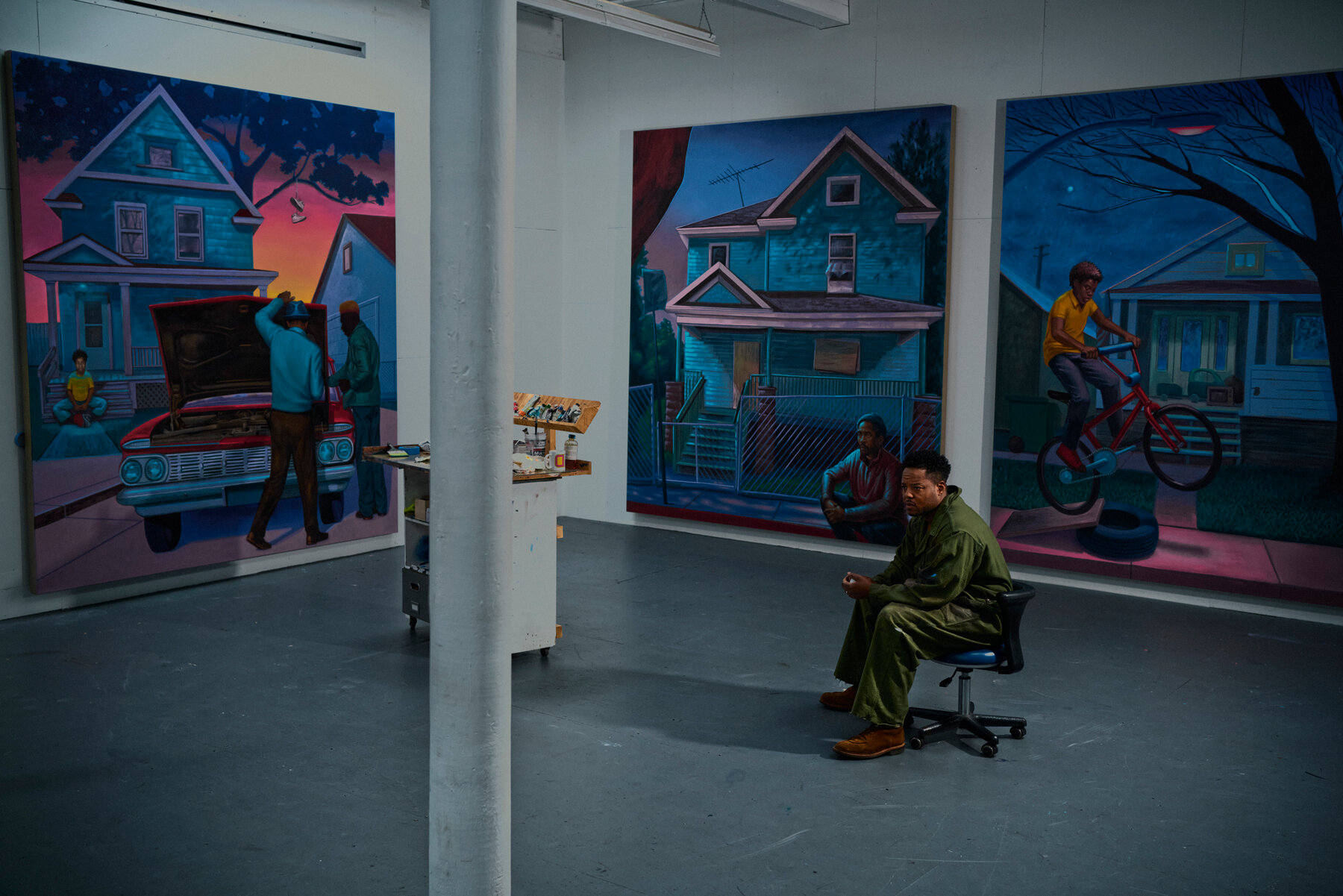 Titus Kaphar sits on an office chair in a gallery space with three large paintings of the exteriors of houses hanging on the walls.