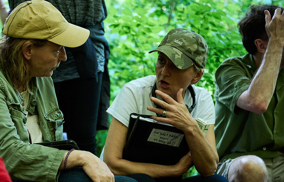 Two women wearing baseball caps sit and talk. One, center, is holding a binder of papers with the label "The Salt Path Draft" on the front.