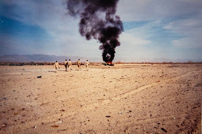 Five special operations troops in desert camouflage walk on barren ground toward a pillar of smoke rising in the distance.