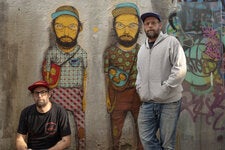 The identical twins Otávio and Gustavo Pandolfo, a.k.a. Osgemeos, with a graffiti of themselves inside their studio in São Paulo. Their painted yellow skin signals their membership in a fantastical world known as Tritrez, part of their “origin story.”