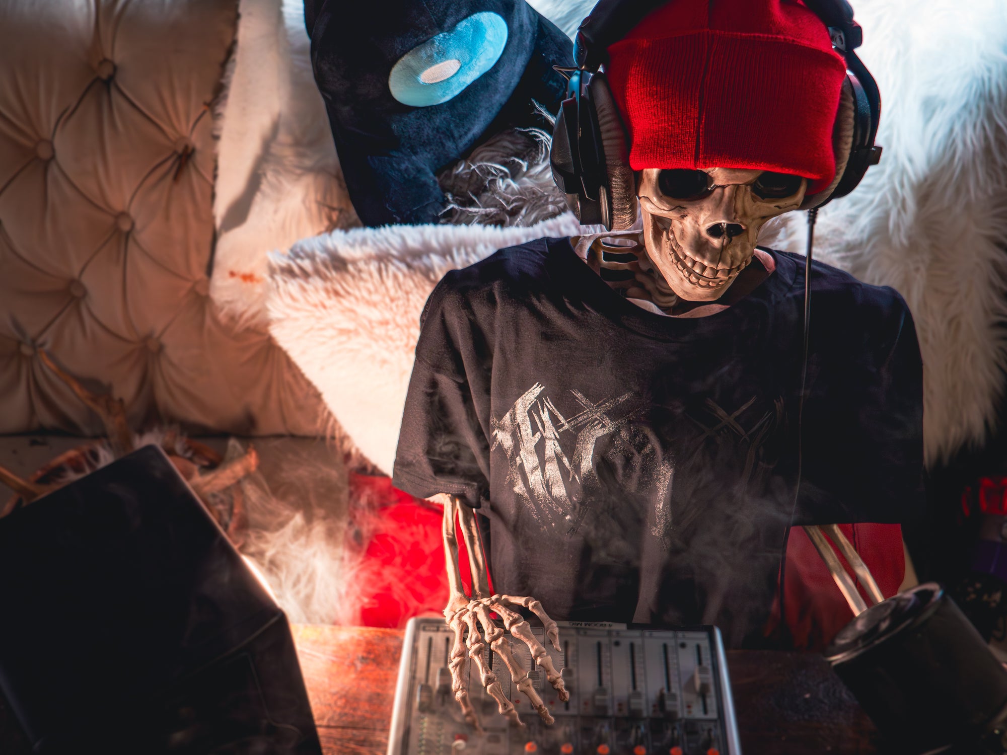 A lifestyle photograph of a skeleton DJing while wearing the Among Us: Black Metal Tee, a Black Crewmate sitting plush is in view of the background behind them.