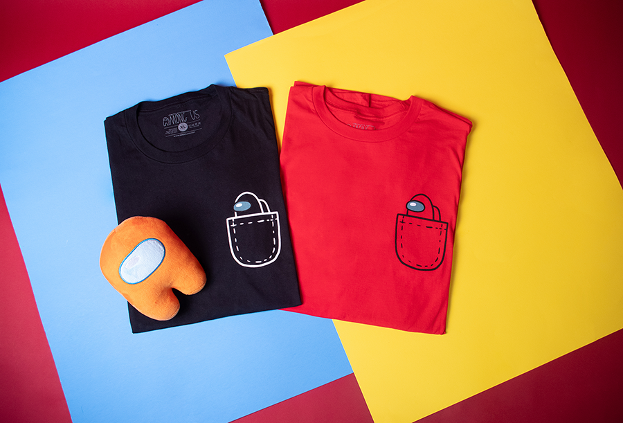 A lifestyle photograph of two Among Us: Mini Crewmate Pocket Tees overlapping side by side. One shirt is in black and the other in Red. An orange frisk wolfie plush rests on top of the black tee next to it.