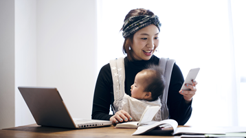 سيدة يابانية مبتسمة تحمل طفلها في حمالة أطفال بينما تفحص هاتفها وتعمل من كمبيوتر محمول