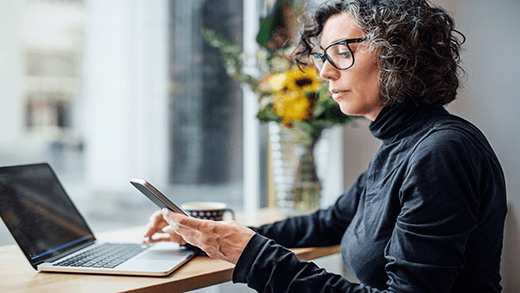 Donna d'affari matura seduta al bar a guardare il suo cellulare mentre lavora al computer portatile. Donna che legge un SMS al bar.