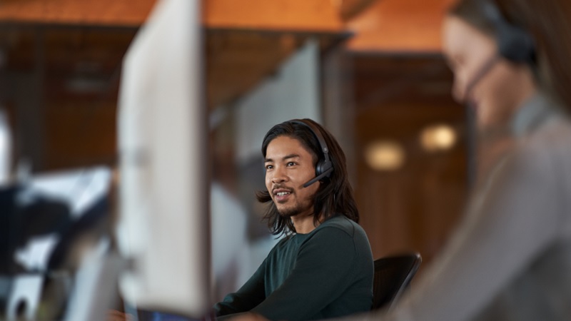 Man met lang haar dat een headset draagt, werkt op een computer in een kantoor, met een collega in de achtergrond