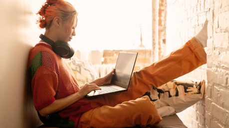 Uma jovem com cabelo laranja senta-se confortavelmente perto de uma janela com fones de ouvido over-the-ear em volta do pescoço olhando para seu laptop Windows 11.