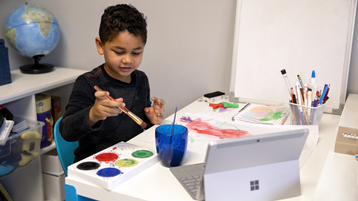 Мальчик рисует красками на бумаге и смотрит в открытый Surface Laptop